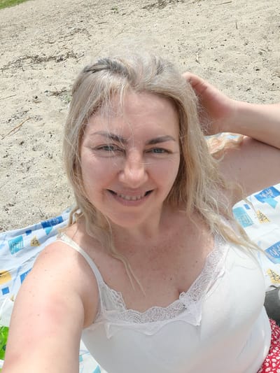 In Sukholuzhzhia, Ukraine, a woman with blonde hair and fair skin with freckles smiles at the camera. She is lying on a beach with sand and scattered debris around her. The woman is wearing a white tank top with lace trim. She has her right arm raised and is touching her hair. A patterned blanket is visible beneath her. The scene appears to be taken outdoors during the daytime.