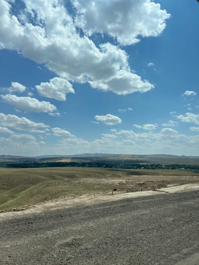 The image depicts a rural landscape under a bright blue sky with scattered white clouds, likely taken in the vicinity of Alqabas, Kazakhstan. The foreground is dominated by a gravel road, appearing somewhat rough and dusty. Beyond the road, a dry, undulating field stretches towards a distant settlement. The field is characterized by sparse, yellowish-brown vegetation, suggesting a dry climate. In the middle ground, a cluster of buildings, possibly a village or small town, is visible, surrounded by patches of green vegetation. The background consists of rolling hills and mountains, fading into a hazy horizon. The overall impression is one of a sun-drenched, arid, and sparsely populated rural area. There are no people visible in the image.