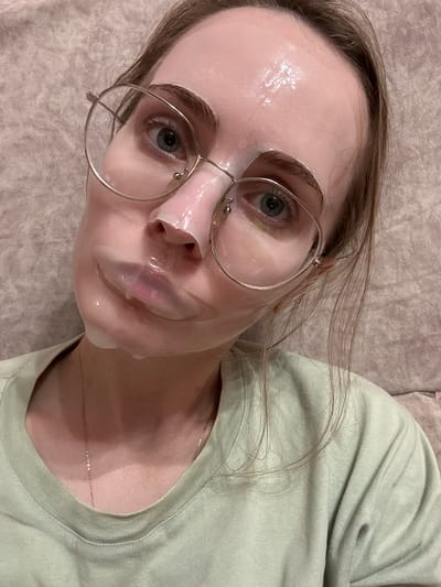 A young adult female is depicted in a close-up portrait, looking directly towards the viewer with a neutral expression. She has light-colored eyes, possibly blue or green, and light brown hair. Her face is covered with a translucent, light pink sheet mask, which appears to be saturated with serum and creates a glossy, moist surface. The mask has cut-outs for her eyes and nostrils, and partially covers her lips. Round, thin-framed metallic eyeglasses are worn over the face mask, resting on her nose and ears. She is wearing a light green, crew-neck top or t-shirt, and a delicate, thin gold-colored chain necklace is visible at her collarbone. The background consists of a soft, textured fabric surface in a light beige or pale pinkish-brown hue, likely upholstery. The lighting is soft and even. The image does not contain identifiable visual information to determine its location as Shirkovo, Russia.