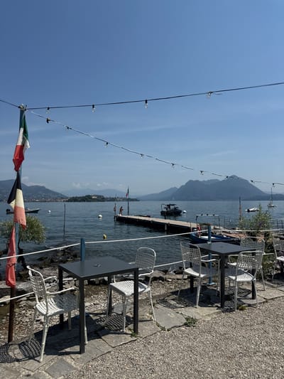 The image depicts an outdoor seating area at a waterfront establishment, likely a cafe or restaurant, with a view of a lake and mountains in the distance. In the foreground, there are two tables, one small square dark gray table with four white metal chairs arranged around it, and another similar table with two chairs visible. The tables and chairs are situated on a stone and gravel patio.
Behind the seating area is a rocky shoreline. The lake is calm and blue, with several buoys visible. A wooden pier extends into the water, with a small boat docked at its end. There are other boats scattered on the lake.
In the midground, on the left side of the image, is a flagpole with an Italian flag and another flag beneath it. A small tree with green foliage is also present. Further in the background, there are buildings on a distant shoreline and rolling mountains under a clear blue sky. String lights are strung across the top of the frame, suggesting the area is set up for evening dining.
The scene appears to be a tranquil, sunny day in Campino, Italy, offering a pleasant atmosphere for patrons to enjoy the lakeside scenery.