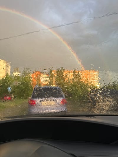 The image captures a view through a rain-streaked car windshield in Volzhsky, Russia. A prominent double rainbow arches across a cloudy sky. In the middle ground, blurred by the rain and distance, is a silver car. Beyond the car, an orange building and green trees are visible. The foreground shows the top of a car's dashboard.