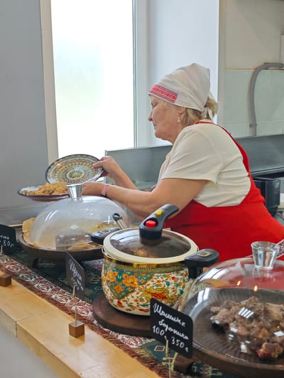 In the city of Cheremushki, Russia, a woman wearing a white headscarf with red trim and a red apron is serving food at a buffet-style counter. She is holding a decorative plate with a colorful geometric pattern and appears to be placing it over a dish. The counter is adorned with a patterned textile and features several food containers. One large pot with a floral design and black handles is prominent. Another dish, covered with a clear dome, contains a rice-like food. Closer to the foreground, a small sign with Cyrillic writing and prices (80, 100, 350) is visible. A glass of water or drink is also present. The background includes a white wall, a window with bright natural light, and some industrial-looking equipment.