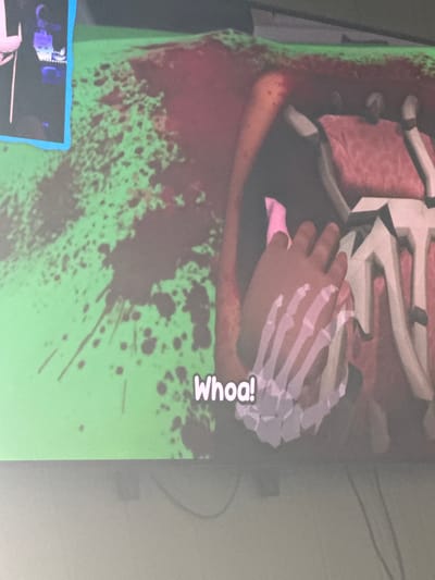 The image displays a screen with a video game scene. In the foreground, a hand with a white, skeletal-like covering on the wrist is visible, reaching towards a pink object. The background features a chaotic environment with green splatters and red, fleshy textures, suggesting a gory or abstract setting. The text "Whoa!" is displayed prominently in white letters near the bottom center of the screen. The image was taken in Moelv, Norway.