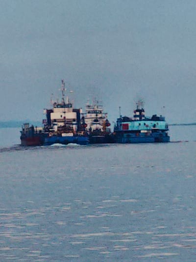 The image shows a calm body of water, likely a river or lake, with two tugboats or workboats moving side-by-side. The sky is overcast and a pale blue-gray, suggesting a cloudy day. The water is a muted blue-gray with some white reflections.
The tugboat on the left is larger and appears to be made of darker colored material, possibly red and dark blue, with a white superstructure. It has several decks and a mast with visible equipment. The tugboat on the right is smaller and brightly painted in turquoise and blue, with a red door on its side. It also has a superstructure with a mast and some visible equipment. Both vessels appear to be moving forward, creating a slight wake behind them.
The overall scene is serene and functional, depicting maritime activity. There are no people clearly visible on the boats. The location appears to be in Russia, given the provided context of Vorogovo. There is no visible text in the image. The time of day is difficult to determine due to the overcast sky, but it appears to be daytime. The weather is mild and uneventful.