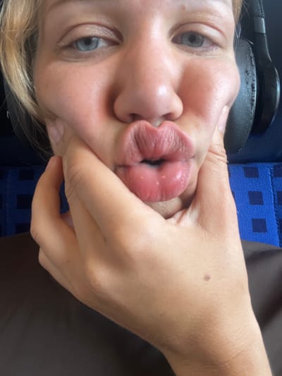 A close-up, frontal shot shows a person's face with blond hair, blue eyes, and plump lips pursed into a kiss. The person's left hand is pushing against their right cheek. The person is wearing black headphones. The background consists of a dark brown shirt and a blue seat with a checkered pattern. The scene does not provide enough information to determine its location in Ampfing, Germany.