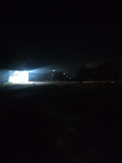 A brightly lit, possibly concrete structure stands in the dark. A beam of light emanates from its left side, creating a visible haze. The surrounding area is dark, with the faint outlines of trees and streetlights visible in the distance. The scene appears to be an outdoor location in Almalyk, Uzbekistan, possibly a street or industrial area at night.