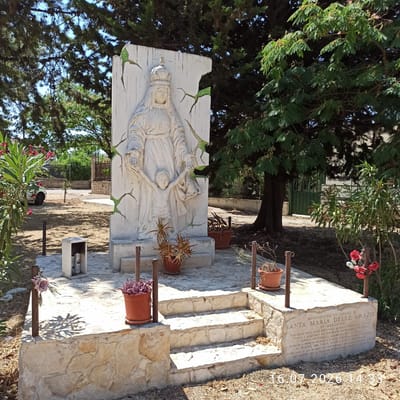 The image shows a stone monument dedicated to "Santa Maria delle Grazie" in Mellitto, Grumo Appula, Italy. The monument features a relief sculpture of a woman, presumably the Virgin Mary, wearing a crown and holding the hand of a child. The sculpture is set against a white, cracked background that appears to be made of plaster or concrete. The monument is situated outdoors in a park-like setting with trees and vegetation. There are steps leading up to the monument, and several potted plants are placed around the base. An inscription on the monument states it was realized by the local committee and dedicated to all mothers and their children. The date "16.07.2026 14:33" is superimposed on the image, suggesting it was taken on this date and time. In the background, a fence, a building, and a green gate are visible. The overall scene conveys a sense of reverence and remembrance.