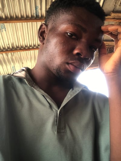 A forensic portrait shot in Aiyetoto-Asogun, Nigeria, depicts a young Black male from the chest up, positioned with a direct gaze into the camera. The subject has short, dark, textured hair and light facial hair growth along his jawline and chin, with minor visible skin texture on his cheeks. He wears a muted olive-green polo shirt featuring a two-button placket, with the top two buttons unfastened, revealing the subtle plaid pattern on the underside of the collar. His left hand is raised, with fingers resting against his forehead above his left eye. The background consists of a corrugated metal roof structure supported by exposed wooden rafters. A bright, undefined light source is visible to the subject's right, creating a backlight effect and illuminating a portion of the background.
