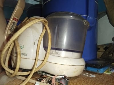 The scene is set indoors on a mottled light brown surface within the Inxuba Yethemba Local Municipality, South Africa. A white Black & Decker food processor forms the central subject, positioned slightly right of center. It features a translucent grey plastic jug attached to its base. The appliance shows signs of use and minor discoloration. A yellowish, coiled power cord rests on and around the processor.
To the immediate left of the processor, partially obscured, is a cream-colored plastic object with a white label bearing a barcode and the text "Cola 0.5L." To the right, a large, bright blue plastic container or bucket stands vertically, partially visible behind the food processor's jug.
In the foreground, to the right of the processor, two electronic devices, resembling mobile phones, are stacked. The top device is dark-colored, and the bottom is light blue. A small, white SIM card tray lies flat on the surface next to them. To the foreground left and center, a crumpled silver-foil packet with red text, partially displaying "RGS," is visible alongside a small packet featuring an image of a woman's face. The overall environment appears cluttered. A light blue wall is visible in the distant background. No individuals are present in the image.