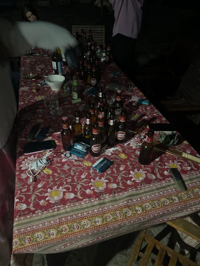 The image depicts an outdoor gathering at night, likely in the city of Felgueiras, Portugal. A long table is spread with a red floral tablecloth. Scattered across the table are numerous bottles of "Super Bock" beer, many of which appear to be empty or partially consumed. Several wine glasses and a white bowl are also visible. Other objects on the table include a mobile phone, a white charging cable and adapter, cigarette packs, and scattered bottle caps. In the background, partially obscured by a white, misty effect, a person with long brown hair is looking downwards. Another person, wearing a light purple top and dark pants, is partially visible on the right side of the frame, facing away from the camera. Wooden chairs are positioned around the table. The overall scene suggests a relaxed social gathering or party.