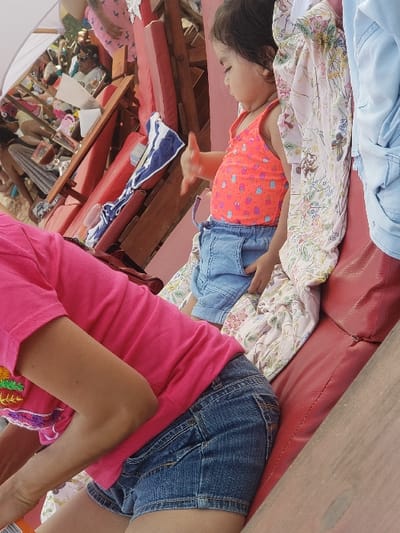 A small child in an orange top and jean shorts lies on a red cushion and floral blanket under a wooden frame. Nearby, an adult in a pink top and denim shorts sits. Other people and items are visible in the background under a white tent.