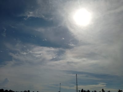 The image displays an outdoor scene under a bright, partly cloudy sky in Meunasahmeucat, Indonesia. The sun, intensely bright and overexposed, is positioned in the upper right quadrant, casting strong illumination over the scene. The sky exhibits a gradient of colors, ranging from a darker blue-grey with scattered wispy white clouds in the upper left to lighter shades of white and grey around the sun, indicating widespread cloud cover. Along the lower horizon, a dark, silhouetted landscape is visible. Two distinct lattice-mast telecommunications towers rise from this horizon: a taller one is located to the right of the frame's center, and a smaller one is positioned further to the left. Beneath these towers, the silhouette reveals numerous palm trees and the outlines of various low-rise structures or buildings. There are no individuals present or engaged in any actions within the visible frame.