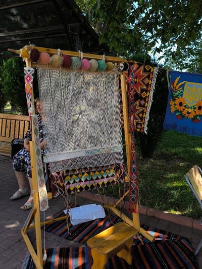 The image depicts an outdoor scene in Illintsi, Ukraine. A large, light-wood upright weaving loom dominates the center, displaying numerous taut white warp threads. A partially woven section near the bottom features horizontal geometric patterns in black, pink, yellow, green, and light blue. Nine yarn balls in varied colors (yellow, maroon, light pink, light green, dark green, dark pink, light blue, purple) are suspended from the loom's top bar. A narrow, vertically oriented woven band with geometric designs hangs from the loom's left support.
To the left of the loom, a light-skinned adult female, dressed in a dark patterned top and light sandals, is seated on a slatted wooden bench, facing forward with a slight smile. She is not actively engaged with the loom. To the right of the loom, a finished black-and-red patterned rug hangs vertically. Further to the right, a blue tapestry depicting the Ukrainian Tryzub emblem surrounded by sunflowers is displayed. A small wooden stool is positioned on a striped woven rug in front of the loom. A stack of white papers or fabric rests on a lower crossbar of the loom. The ground is paved with red-brown interlocking bricks. Green trees and foliage are visible in the background under a blue sky, with a dark metal pergola structure partially visible in the upper left. The scene is illuminated by bright natural daylight.