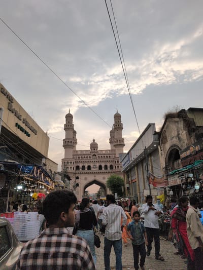 This image captures a bustling street scene in Hyderabad, India, centered on the Charminar monument in the background. The sky is largely overcast with diffused clouds, suggesting a cloudy day or twilight. Multiple dark utility wires crisscross the upper part of the frame.

The Charminar, a light-colored historical structure, dominates the background, featuring four prominent minarets with domes and a large central arched gateway. A clock face is visible on its lower section. Small, dark specks, likely birds, are scattered across the sky near the monument.

On the left side of the street, a building displays the sign "HOTEL ROYAL SWAGATHAM FOREVER." Below this structure, a market stall, covered by a blue tarp, is illuminated by strung lights, showcasing various items. A portion of a silver vehicle is visible in the lower left foreground.

On the right side of the street, a row of commercial buildings includes an older, weathered structure with an arched entrance and a more modern building with roll-down shutters. A "XEROX" sign is visible on a shop facade. Market stalls displaying merchandise extend along this side of the street.

The street itself is densely populated with pedestrians walking in various directions. In the immediate foreground, facing slightly right and away from the camera, is a person wearing a brown and white plaid shirt. Further back, a man in a white shirt and dark pants walks alongside a young boy in a blue shirt and dark pants in the right-center midground. Other individuals are distributed throughout the scene, engaged in pedestrian movement and shopping activities. The ground appears to be paved with stones or cobblestones.