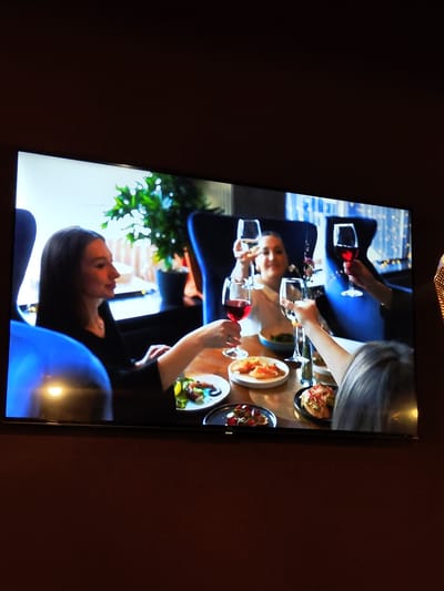 The image shows a television screen displaying a scene of people dining. In the foreground, a woman in a black top is seated at a table, holding a glass of red wine. In the background, three other individuals are visible, also holding wine glasses. The table is set with several plates of food, including appetizers. The environment appears to be a restaurant setting, with plush seating and decorative lights. The television is mounted on a dark brown wall. The image does not contain any information that would indicate a specific location in Zhukovsky, Russia.