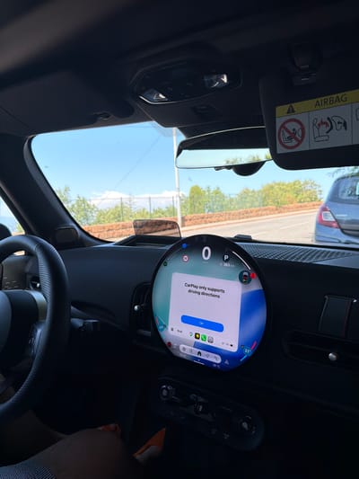 This is a first-person view from inside a car, looking at the dashboard and the road ahead. The car appears to be moving on a sunny day, as indicated by the bright sky and the shadows. The dashboard features a large circular digital display showing speed, parking gear, and distance to destination. A pop-up message on the screen reads "CarPlay only supports driving directions." Below the display are climate control buttons. To the right, a sun visor with an airbag warning sticker is visible. Outside the car, a road is visible with trees and a brick wall on the side. Another car, a grey hatchback, is driving ahead. The location is identified as Théoule-sur-Mer, France, likely on a coastal road given the vegetation and the distant sea visible through the windshield.