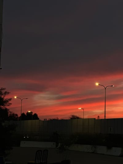 The image depicts an outdoor scene in Kfar Vitkin, Israel, during twilight or sunset. The sky, occupying the upper two-thirds of the frame, features prominent horizontal strata of vibrant red and orange clouds near the horizon, transitioning to a dark grey and almost black hue overhead. Four streetlights are illuminated, emitting warm yellow light: a double-headed pole stands prominently on the right, a single-headed pole on the left, and two smaller, single-headed poles are visible further in the midground. A tall, dark, continuous barrier, likely a sound wall, extends horizontally across the midground, behind which the dark silhouettes of trees are visible against the illuminated sky. In the immediate foreground, two dark, upright chairs with slatted backs are positioned on a dark, paved surface. Two light-colored concrete barriers are situated between the chairs and the main barrier. A dark, vertical structural element is partially visible along the extreme left edge of the image. No individuals or discernable activities are present.
