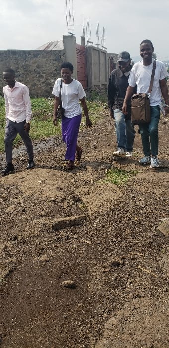 The image shows four individuals walking on an unpaved dirt path in Buzove, Democratic Republic of the Congo, under an overcast sky. From left to right: a male in a light-colored collared shirt, dark trousers, and dark shoes; a female in a white t-shirt, purple trousers, and sandals, carrying a dark shoulder bag; a male in a dark shirt, blue jeans, light athletic shoes, and a dark baseball cap; and another male in a white t-shirt, blue jeans, and light athletic shoes, carrying a brown shoulder bag and holding papers in his left hand. The female and the rightmost male are smiling. The ground is composed of loose brown soil, uneven with slight mounds and depressions, and contains sparse green vegetation. In the background, a low stone wall, a red-brown metal gate, and unfinished concrete structures with exposed rebar are visible. Distant terrain or structures are faintly discernible on the horizon to the far right.