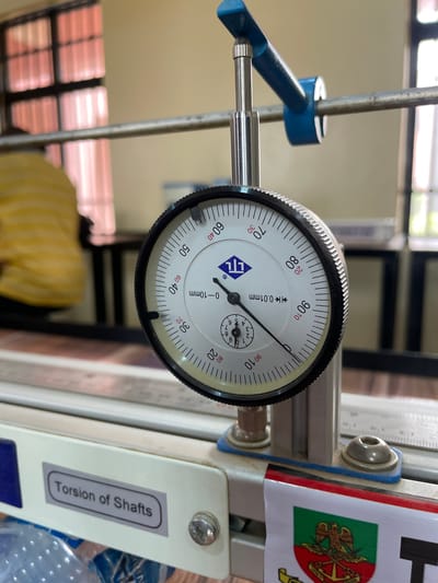 The image shows a close-up of a dial indicator gauge, which is a precision measuring instrument. The needle on the main dial points between "80" and "90". The smaller dial indicates a value around "5". The dial indicator is attached to a metal frame, and a thin metal rod extends from the gauge, presumably to make contact with the object being measured. A label on the frame reads "Torsion of Shafts". A flag with a heraldic emblem is visible on the frame. The dial indicator is placed somewhere in Afaka, Nigeria.