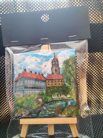 This is a close-up, eye-level shot of a small, framed painting resting on a miniature wooden easel. The painting depicts a picturesque scene of a European-style castle or palace complex. The architecture features a light blue building with red-tiled roofs on the left, and a taller, stone tower with a dark spire on the right. Lush green trees surround the buildings, and a body of water, possibly a river or canal, flows in the foreground. The sky is a vibrant blue with fluffy white clouds, suggesting pleasant weather.

The painting is encased in clear plastic packaging, which is attached to a black cardstock backing. The backing has a circular cutout at the top, likely for hanging, and two small white slits. The overall presentation suggests it is a souvenir or decorative item. The background of the image is a textured, possibly reflective surface with a pattern of circular perforations, creating a bokeh effect. No people are visible in the painting or the surrounding context. The image appears to be taken indoors, possibly in a gift shop or at a market stall in České Budějovice, Czechia, given the provided location context. There is no visible text in the image.