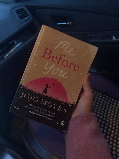 The image depicts a human hand holding a paperback book titled "Me Before You" by Jojo Moyes inside what appears to be a vehicle. The specific location in Apshak, Uzbekistan, cannot be determined from the visual information provided.
The book has a light brown cover with the title "Me Before You" in script, primarily red text. Below the title, a silhouette of a woman with outstretched arms and a small bird is visible against a pink and dark red gradient background, above a dark foreground depicting foliage. The author's name, "JOJO MOYES," is printed on a black strip at the bottom of the cover, along with a critical review quote and the Penguin Books logo.
A right human hand, with light skin tone, holds the book on its right edge. The person is wearing a fuzzy, light pink or purple sleeve garment. In the background, to the left, a dark vehicle interior is visible, including an air vent and part of a dashboard or door panel. To the right, a dark, textured surface, possibly a seat cover with a raised grid pattern, is present. The overall lighting is subdued.