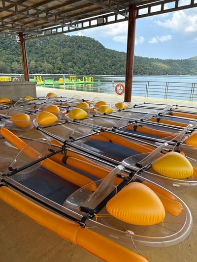 The image shows a row of kayaks, likely stored at a rental facility, in Phu Quoc, Vietnam. The kayaks are clear, hard-shelled models with orange inflatable pontoons on either side and yellow buoys attached to the front. They are racked against a wall. In the background, visible through the structure's framing, is a view of the coastline, with a tree-covered hill and a body of water, presumed to be the sea. The picture is taken from under a covered structure, possibly a loading dock or shelter. A life preserver hangs on a railing. The overall scene suggests a recreational area or water sports rental location.