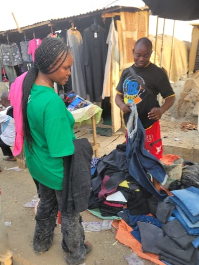 Two people are browsing a pile of clothes in an outdoor market. A man in a black shirt with a logo is holding up a pair of jeans. A woman in a green shirt is looking at the clothes; a pink scarf is draped around her. The clothes are piled on the ground, and include jeans, shirts, and other garments. The market is bustling, with other shops and people visible in the background. The ground appears to be dirt.