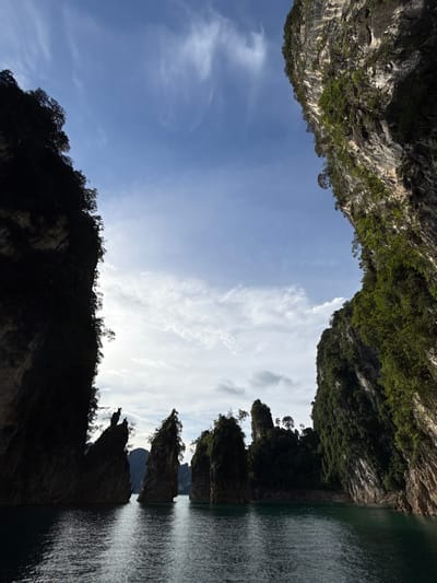 This image depicts a serene natural landscape with a clear blue sky and scattered white clouds. The foreground is dominated by dark, silhouetted rock formations and lush green trees, suggesting a mountainous or jungle terrain. The formations are sharp and jagged, with some trees growing precariously on their edges. To the right, a body of calm, dark water reflects the surrounding scenery. The water appears placid, with gentle ripples on its surface.
The composition suggests a view from a boat on the water, looking up towards the sky and the surrounding cliffs. The orientation of the image is unusual, with the sky appearing at the bottom and the water at the right, indicating the photo may have been taken vertically and rotated. The lighting suggests it is daytime, possibly late morning or early afternoon, given the brightness of the sky and the contrast between light and shadow. There is no visible text in the image. The overall mood is one of natural beauty and tranquility, with the imposing rock formations contrasting with the peaceful water and sky. The location cues, such as the karst-like rock formations and the tropical foliage, are consistent with a location like Khao Phang, Thailand, known for its stunning limestone cliffs and emerald waters.