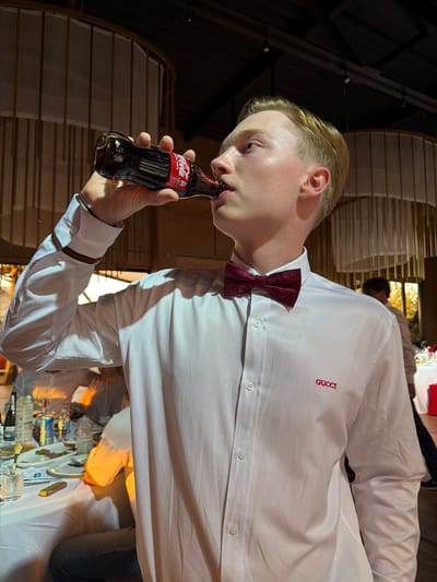 The image shows a young man with blond hair wearing a white dress shirt with a red Gucci logo on the left breast and a burgundy patterned bow tie. He is drinking from a small glass bottle of Coca-Cola. The man is at a formal event, possibly a wedding or a gala, given the white tablecloths and table settings in the background. The lighting suggests it's indoors, and likely evening. The scene appears to be set in a banquet hall or restaurant, with decorative elements like layered screens visible behind him. Another person, partially obscured, is also visible in the background. The man's expression is neutral as he sips his drink. The Coca-Cola bottle has visible text that includes "Coca-Cola" and "Original Taste".