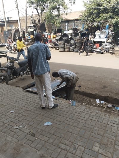 The image depicts a daytime street scene in Kofar Mazugal, Nigeria. In the immediate foreground, a grey block-paved sidewalk, littered with plastic and paper debris, borders an unpaved earthen drainage ditch. Two adult males are central figures: one, viewed from behind, wears a light blue and white checkered short-sleeved shirt, light-colored trousers, and dark shoes, standing upright on the sidewalk. To his right, a second adult male, in a light-colored long-sleeved shirt and dark trousers, is bent over a rectangular metal object placed within the ditch, actively engaged in work; two small paint cans are visible on the ground nearby.
Beyond the ditch, an unpaved road stretches into the midground. A black motorcycle is partially visible, parked to the left. Two additional adult males are present on the road: one, wearing a yellow hooded top and blue jeans, walks from left to right, while another, dressed in dark clothing, walks across the road in the same direction. The background features a dense outdoor market or workshop specializing in metal goods. Numerous items are displayed or stacked, including large dark metal drums, satellite dishes, industrial-sized pots, and various metal frameworks and furniture. Buildings, green foliage from trees, and overhead utility lines are visible in the distant background. The scene is illuminated by bright natural light.
