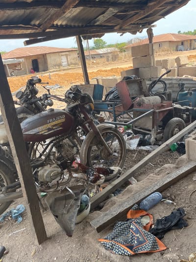 A cluttered scene features a dilapidated motorcycle with a "FraJeno" inscription. It is surrounded by various discarded objects including wooden planks, a tire, scraps of fabric, and parts of machinery. In the background, cinder blocks are visible along with simple buildings. The scene is under a partial roof of corrugated metal, suggesting an outdoor storage area or makeshift workshop. The overall impression is one of disarray and neglect.