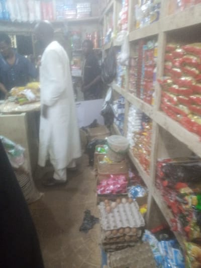 The image depicts the interior of a small grocery store, likely a local market stall, situated in Tundun Wada South, Nigeria. The scene is somewhat cluttered, with shelves packed with various packaged goods, predominantly food items. Prominently displayed are rows of snacks or biscuits in red and yellow packaging, suggesting a focus on confectionery or convenience foods. On a lower shelf, a carton of eggs is visible, with several eggs already placed in their individual compartments. To the left, a person dressed in a white robe stands, appearing to be either a customer or the shopkeeper. Another individual in a dark shirt is partially visible in the background, also seemingly within the store. The lighting is dim, contributing to the overall blurry and somewhat indistinct quality of the image. The environment suggests a typical informal retail setting, common in many Nigerian urban or semi-urban areas.