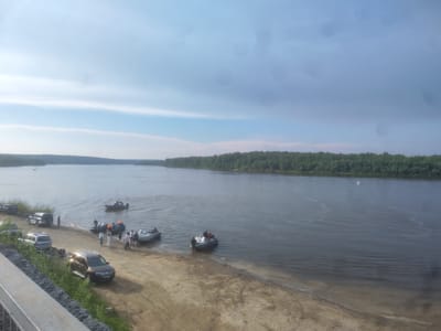 The image depicts a wide, calm river with a tree-lined bank on the opposite side. The foreground shows a sandy shore with several vehicles parked, including SUVs and a station wagon. A group of people, dressed in casual clothing and life vests, are gathered near several motorboats on the water's edge. Some boats are in the water, while others are partially on the shore. The sky is overcast with some patches of blue. A small white sailboat is visible in the distance on the river. The scene suggests a recreational or fishing activity.