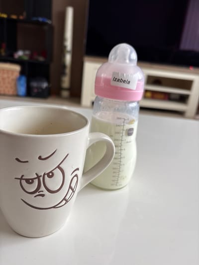 The image shows a close-up of a white mug with a cartoonish, mischievous face printed on it. The mug is half-full of a light-colored liquid. Next to the mug is a baby bottle with a pink nipple and collar, also containing a light-colored liquid. A white label with the name "Izabela" is attached to the bottle. The bottle has measurement markings in milliliters. Both the mug and bottle are placed on a white, reflective surface, likely a table. In the blurred background, a dark television screen and a white shelving unit are visible. The scene appears to be indoors, in a domestic setting. There is no indication of the location in the city of Veghel, Netherlands.