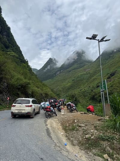 A line of parked motorcycles and a white SUV, a Honda BR-V, are on the side of a winding road in Yên Minh Commune, Vietnam. The road is next to a steep, green, mountainous terrain partially obscured by clouds. A person in a red jacket stands on the edge of the road. A tall metal pole with solar panels and streetlights is visible.