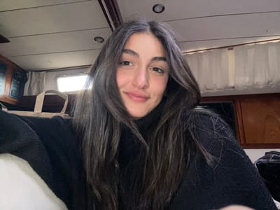 A young adult female with long, dark brown hair, parted down the middle, is depicted from the chest up, looking directly at the camera with a slight smile. She wears a black ribbed turtleneck sweater. The scene takes place within the interior cabin of a boat, likely docked in La Grande-Motte, France. The background shows a light-colored ceiling with recessed lighting and a dark wooden beam running horizontally. To the left, a small rectangular window (portlight) is visible with bright light outside, along with a portion of a wooden wall featuring what appears to be a control panel with dark readouts. A light-colored canvas bag rests on a surface beneath the window. To the right, another section of wooden paneling and white curtains covering another window are visible. The overall lighting is natural from the windows, supplemented by the boat's interior lights.