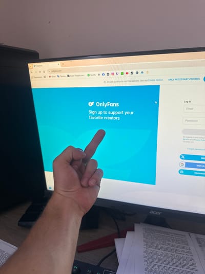 An indoor personal workspace, identified as being located in Saraia, Bulgaria, is depicted from a first-person perspective. In the foreground, a light-skinned forearm and hand are prominently positioned, extending from the lower right towards the center of the frame. The hand is engaged in an action, displaying a middle finger gesture directly towards a computer monitor.
The central subject is a black Acer-branded computer monitor, displaying a web browser interface open to "onlyfans.com". The webpage features a blue background with the white "OnlyFans" logo and text stating, "Sign up to support your favorite creators." To the right, a login panel includes fields for "Email" and "Password," alongside "Log in" options via X (formerly Twitter), G (Google), and a password manager icon. A cookie notice is visible across the top of the main content area, stating, "We use cookies to run this website. See our Cookie Notice." The browser's top bar displays a tab for "OnlyFans" and various bookmarks, including "Приложений" (Applications), "M Gmail," "Перевод" (Translate), "Apple TVapple.com," "Spotify," and icons representing social media platforms such as Facebook, Instagram, Twitch, TikTok, YouTube, and Binance.
The monitor rests on a dark wooden desk. To the right of the monitor and in the foreground, multiple sheets of white paper with black text, appearing to be in Cyrillic script, are stacked. A black keyboard with some green keycaps (F4, F5, F6 visible) is partially visible at the bottom left. A dark, rectangular electronic device, possibly a smartphone or external drive, lies on the desk to the left of the visible hand. The background reveals a plain, light-colored wall above the monitor. The overall lighting is even, suggesting an adequately lit room.