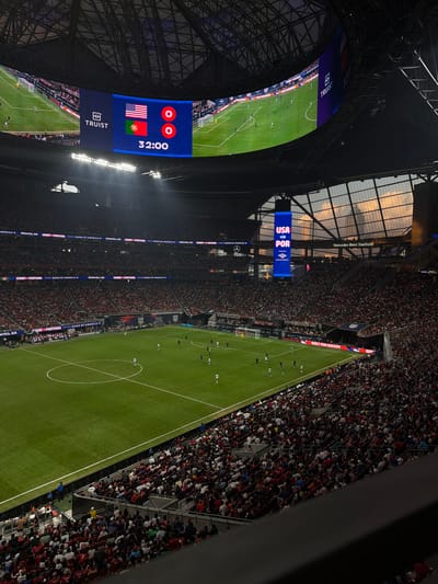 The image captures a soccer game between the USMNT and Portugal at the Mercedes-Benz Stadium in Atlanta, GA. The stadium is filled with spectators. The score shows a 0-0 tie at the 32:00 mark. A large screen displays the game action. Sponsorships such as Truist and Bank of America are visible.