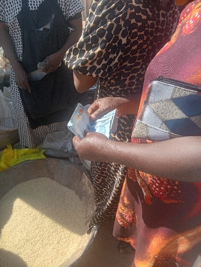 In the city of Gurku Gwari, Nigeria, an outdoor market scene depicts a transaction involving three individuals. The central figure, wearing a black and brown leopard-print garment, holds and counts several light-blue Nigerian 1000 Naira banknotes with both hands. To their left, another individual, dressed in a black and white patterned top and a black apron, has their hands near the apron's front pocket. To the right, a third individual, partially visible, wears a colorful garment with red, orange, and white patterns, and a grey and blue patterned purse rests against their side. In the foreground, a large, weathered metallic basin contains a significant quantity of light-yellow granular material, consistent with rice or millet. A yellow fabric or plastic sheet is present on the ground beside the basin. White sacks are visible in the background.
