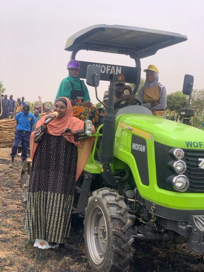 A woman wearing a black and white dress and a peach headscarf is standing in front of a green tractor, holding a microphone and speaking to an audience. She is standing in a field of dirt and grass, and the tractor is parked in front of a group of people. The tractor is green and has a black canopy. The woman is the only person in the foreground, and the others are in the background. The tractor is a WOFAN model. The scene is in the city of Ringimawa, Nigeria, at the coordinates 11.666 latitude and 8.416 longitude.