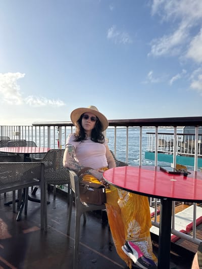 A woman sits at a red table on a ferry or boat under a blue sky with clouds. She wears a light pink top, a straw hat, and sunglasses. Her left arm has tattoos. A yellow patterned cloth rests on her lap. She is wearing white, pink, and grey sneakers. A brown purse sits next to her on the chair. The ferry has metal railings and perforated metal chairs. The water can be seen beyond the railings. The location is likely Corralejo, Spain based on the surroundings.