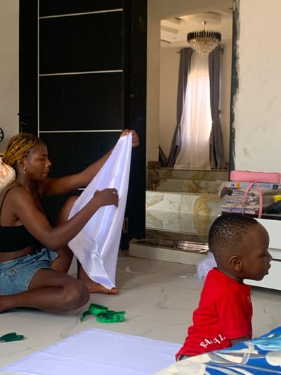 An indoor residential scene, presumably located in Achalla, Nigeria, features two individuals: an adult female and a young child. The adult female, with light brown braided hair, is seated barefoot on a light-colored tiled floor, wearing a black crop top and blue denim shorts. She is actively handling a piece of white fabric, holding it vertically with both hands, suggesting a task involving textile manipulation. A green measuring tape and a pair of green-handled scissors are on the floor near her right side, next to a larger piece of white fabric spread flat. To the right, a young male child, with short dark hair and wearing a red t-shirt, is seated on the floor, facing away from the camera and the woman. His gaze is directed to the right.
The background reveals a dark doorway or doorframe behind the woman, leading to a multi-level interior. Beyond this doorway, a staircase with light-colored patterned tiles ascends, visible through an archway. At the top of the stairs, a large window is draped with both sheer white and heavy grey curtains tied back. A multi-tiered chandelier with dark metal and crystal elements hangs from the ceiling above the stairs. To the far right, partially obscured, a white storage unit or shelf holds several stacked books and papers secured with string, topped by a pink item. The room is brightly lit, likely by natural light from the window. The overall environment suggests a domestic setting where fabric-related work is being conducted.