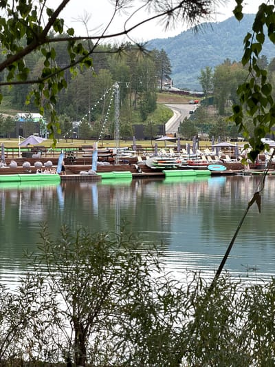 This is an outdoor scene at a lake with a wooden pier. Several people are visible on the pier, some sitting in chairs, others standing. There are various objects on the pier, including lounge chairs, umbrellas, and paddleboards or kayaks lined up. Green safety barriers are positioned along the edge of the pier. In the background, there are trees and a forested hillside, with a road and some vehicles visible on it. A tall white metal structure with triangular flags is also present, suggesting an event or competition might be taking place. The water reflects the sky and surrounding elements, indicating it is likely daytime, and the overcast sky suggests cloudy or mild weather. The overall impression is of a recreational area by a lake, possibly during a public gathering or a sporting event. Text visible on blue banners on the pier appears to read "ЦЕНТР ПЕДАГОГИЧЕСКОЙ АКТИВНОСТИ".