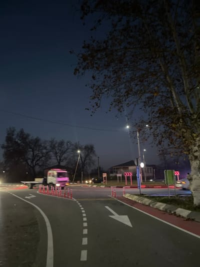 This image captures a road roundabout at dusk or night in the city of ლესის თემი, Georgia. The sky is dark blue, illuminated by artificial light sources. The asphalt road features white dashed and solid lane markings, including white directional arrows. A dedicated red-painted lane, likely a bicycle path, is visible on the right, separated from the main road by a raised curb and a line of orange and white reflective bollards.
A white flatbed truck, illuminated by a pink light source and with its headlights on, is transiting through the left side of the roundabout. The roundabout's central island is delineated by additional orange and white reflective bollards and multiple red and white chevron signs pointing right. Several streetlights illuminate the intersection, and the headlights of other distant vehicles are visible on the left side of the frame. Deciduous trees with sparse, brownish leaves are present on the right and in the background, alongside a residential-style building partially visible beyond the roundabout.