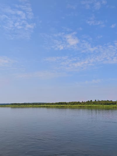 The image shows a wide, calm river with a dense line of green trees and vegetation along the far bank. The sky above is a clear light blue with scattered white, wispy clouds. The water reflects the sky and the distant treeline, creating a peaceful and natural landscape. The photograph appears to be taken from a boat on the river, looking towards the shore. The location is identified as Novonazimovo, Russia, a village situated on the bank of the Angara River.