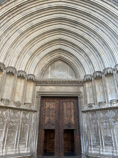 The image shows the grand entrance of the Girona Cathedral in Girona, Spain. The entrance features a large set of ornate wooden double doors, with the left door slightly ajar, revealing a glimpse of the interior. Above the doors, a series of progressively smaller arches rise towards the apex of the facade, creating a dramatic architectural effect. The stonework is intricate, with detailed carvings and decorative elements adorning the facade. The lighting suggests it is daytime, casting subtle shadows that highlight the depth and texture of the stone. No people are visible in the frame.