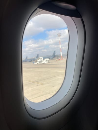 The image shows an airplane window with a view of an airport tarmac on a partly cloudy day. In the distance, two airplanes are visible, along with a set of mobile stairs parked near one of them. A tall, red and white striped tower, likely an airport marker or antenna, stands prominently on the right side of the frame. The tarmac is a paved surface with yellow markings. The foreground is dominated by the dark interior of the airplane's cabin, framing the exterior view. The location is identified as the city of Yagel, Israel.