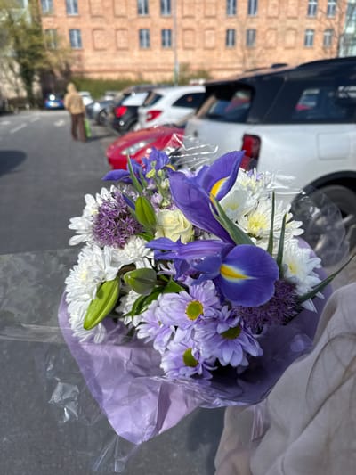 The foreground of the image features a floral bouquet held by a person's arm, visible in the lower right. The bouquet is composed of purple irises with yellow centers, white chrysanthemums, light purple daisies, and spherical purple allium blossoms, wrapped in light purple paper and clear cellophane. The background, out of focus, reveals an outdoor parking area within the city of Wyre Forest, United Kingdom. Multiple vehicles are parked, including a white SUV on the right and a red sports car further left. A lone individual, wearing a light brown hooded jacket and dark trousers, stands facing away from the viewer in the left midground, near a blue car. A multi-story brick building with numerous windows occupies the distant background. The scene is illuminated by daylight, casting shadows.