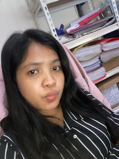 A person lies on a light pink surface, likely bedding. The person has dark hair and is wearing a black and white striped shirt. Adjacent to the bedding, a metal shelf is visible, filled with a collection of files and folders. The location is Negara, Indonesia.