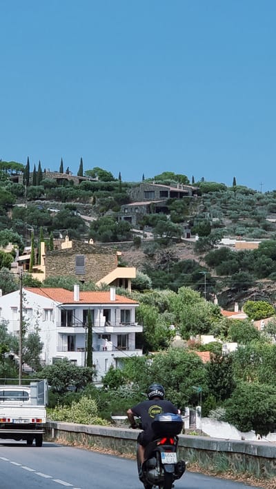 The image depicts a hillside residential area in Cadaqués, Spain, with a clear blue sky overhead. In the foreground, a person is riding a black scooter on a paved road. The scooter has a license plate reading "6645 LJL". To the left of the scooter, a white truck with a license plate reading "7767 MNS" is visible.
The hillside is covered with numerous houses of varying architectural styles, many constructed with stone and featuring red-tiled roofs. These homes are interspersed with dense green vegetation, primarily olive trees, and scattered cypress trees. On the upper slopes, larger, modern houses with stone facades are prominent. In the midground, a white multi-story building with balconies and red-tiled roofs is partially obscured by trees. Further up the hill, a lighter-colored building with yellow accents is visible. The overall impression is of a well-established, affluent residential community integrated into the natural landscape.